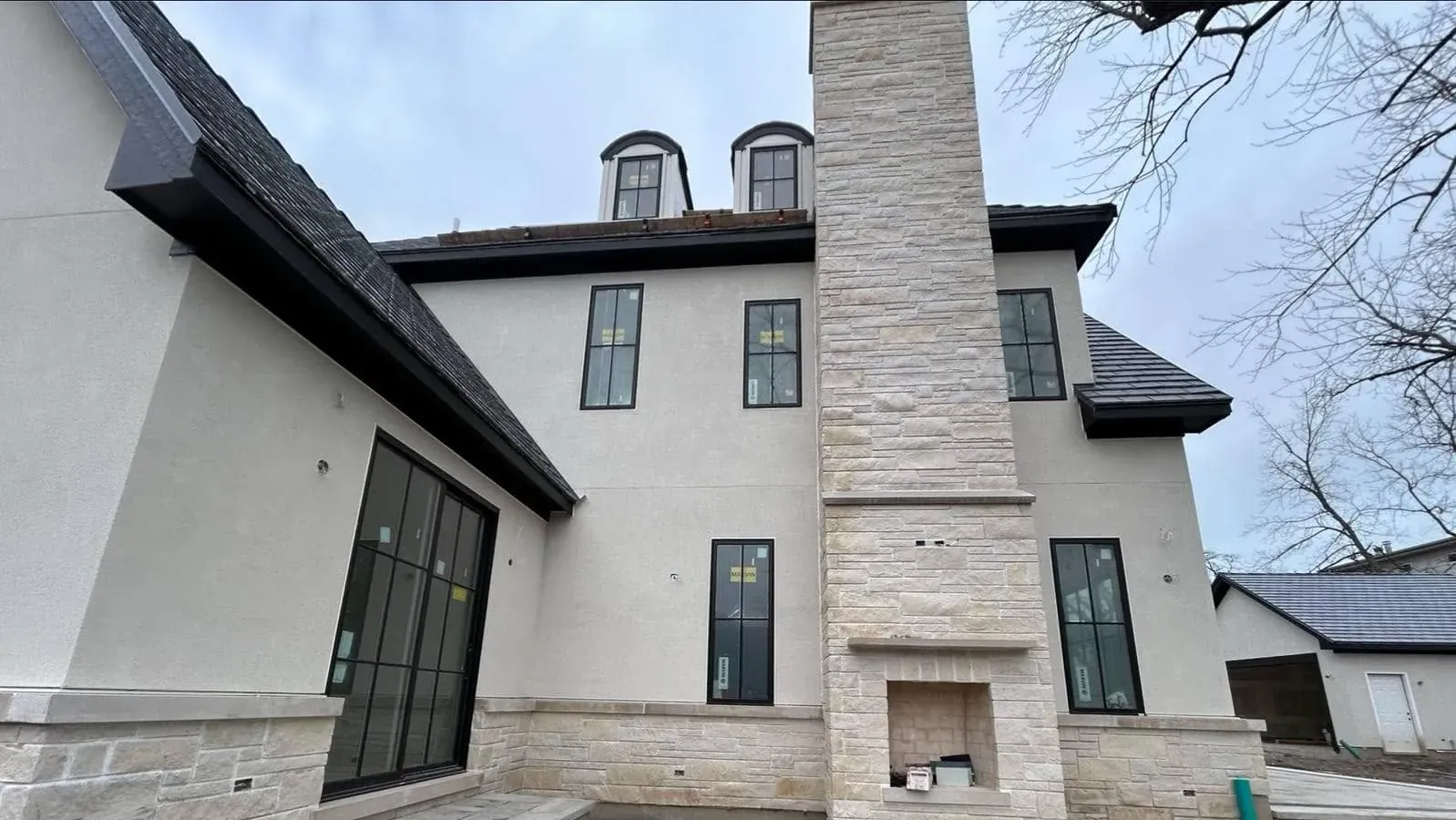 Finished rear elevation of a custom home — a smooth white stucco field meeting a full-height coursed natural stone chimney with an outdoor fireplace, under dormers and tall black-framed windows