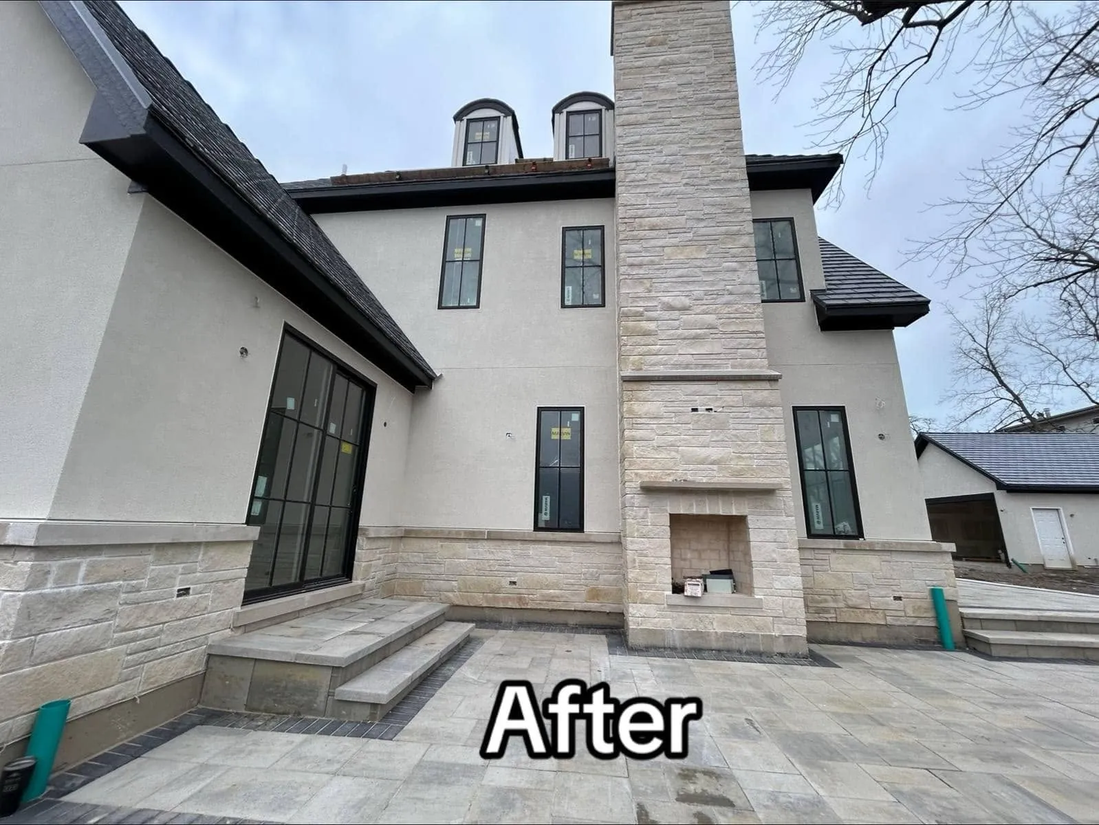 Finished rear elevation of a custom home with a smooth white stucco field, a full-height coursed natural stone chimney housing an outdoor fireplace, dormers and a paver patio