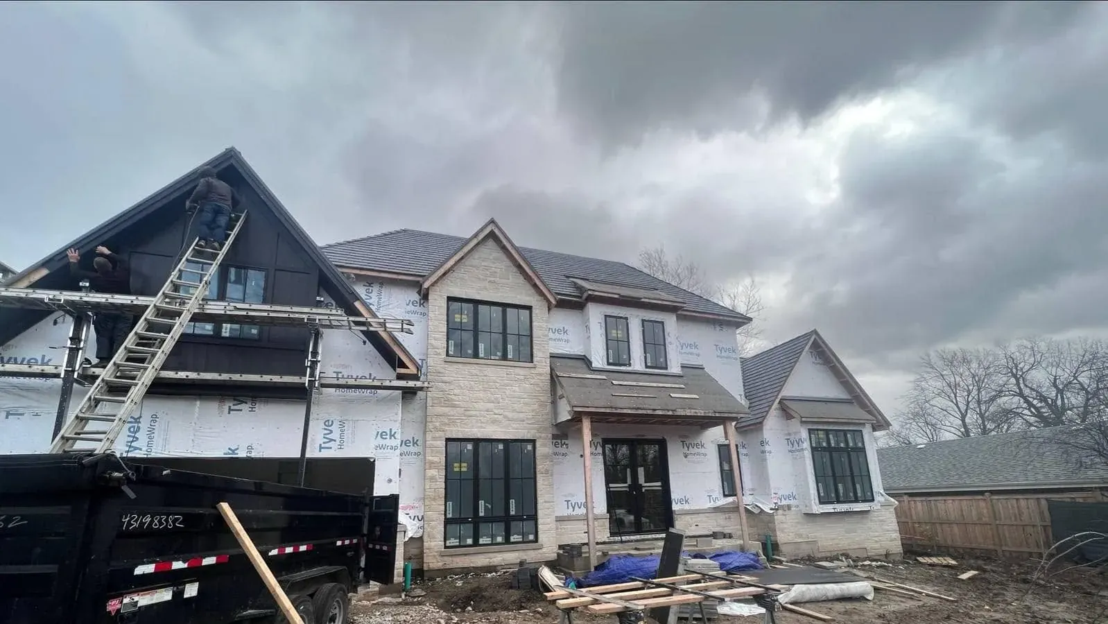 Custom home mid-build with Tyvek HomeWrap over the sheathing, a worker on a ladder at the front gable and stone veneer started on the right wing