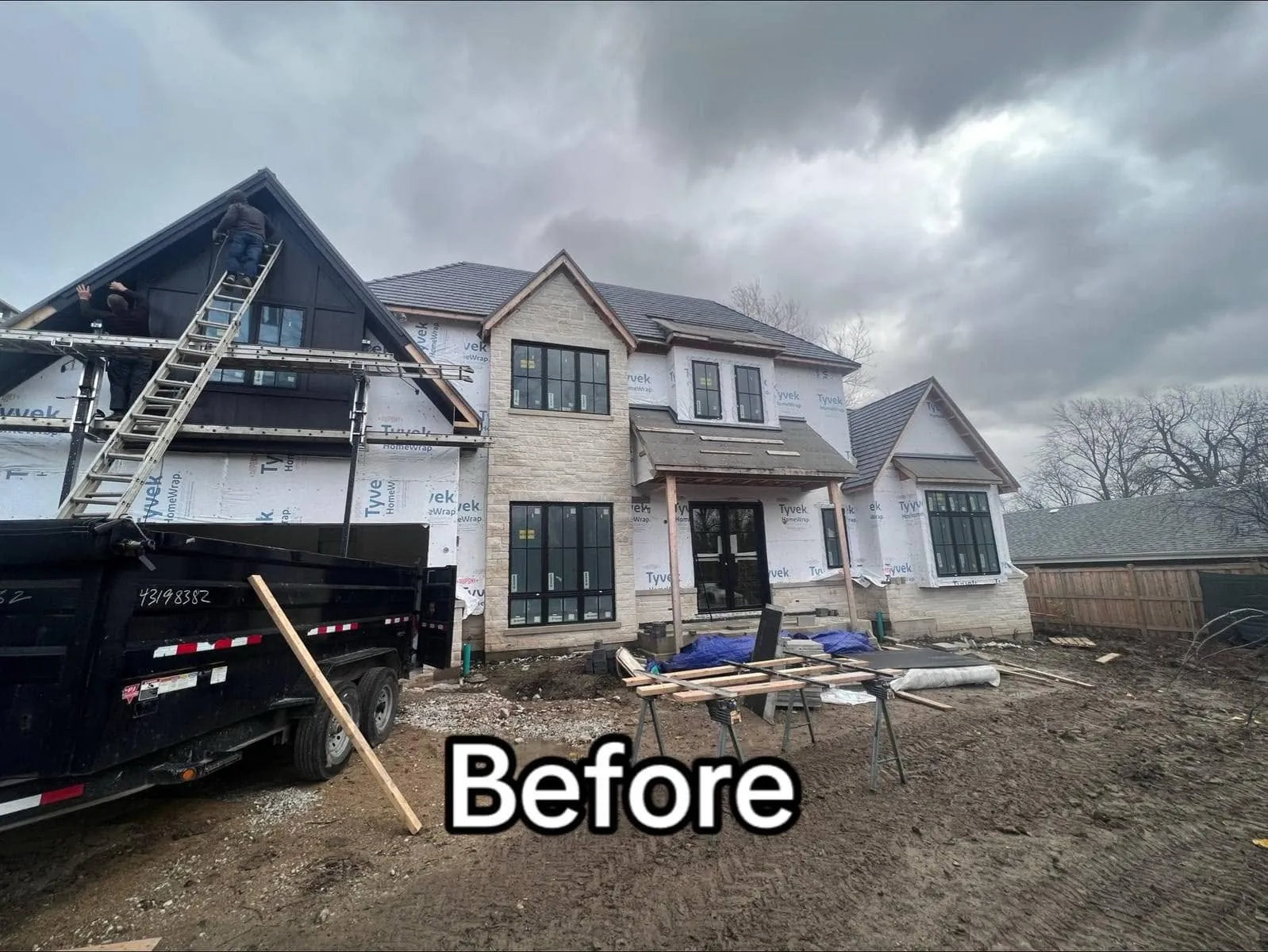 Custom home mid-build with Tyvek HomeWrap over the sheathing, a ladder against the front gable and stone veneer started on the right wing
