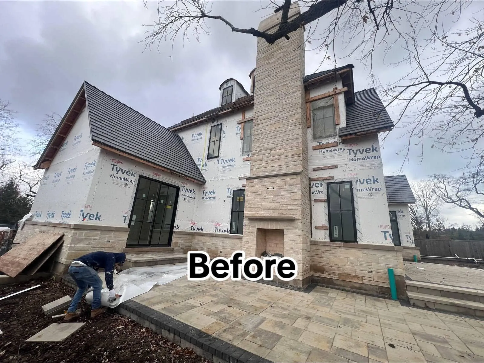 The same custom home's rear elevation mid-build, wrapped in Tyvek HomeWrap, with the natural stone chimney already built and the paver patio laid